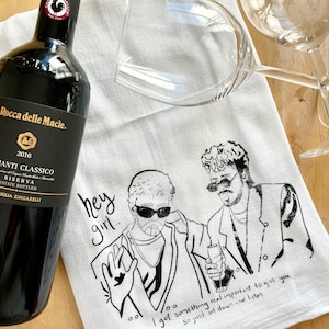 May include: A bottle of Rocca delle Macie Chianti Classico wine next to a white tea towel with a black and white illustration of two men. The towel has the text "hey girl" and "I got something real important to give you". Two wine glasses are also in the image.