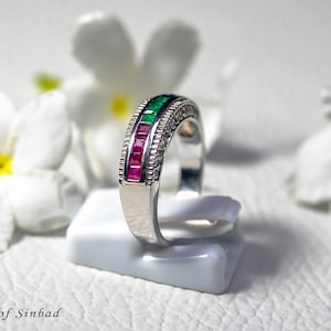 May include: A silver ring with a row of green and red gemstones, and a row of small clear stones. The ring is displayed on a white stand, with white flowers in the background. The text "Treasures of Sinbad" is at the bottom.