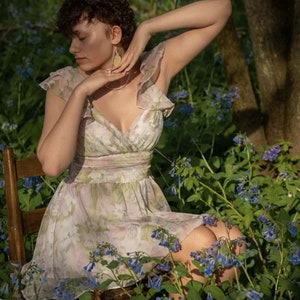 May include: A floral print dress with a V-neck and ruffled sleeves. The dress is light-colored with a pink and green floral pattern. The person is sitting on a wooden chair surrounded by blue flowers and greenery.