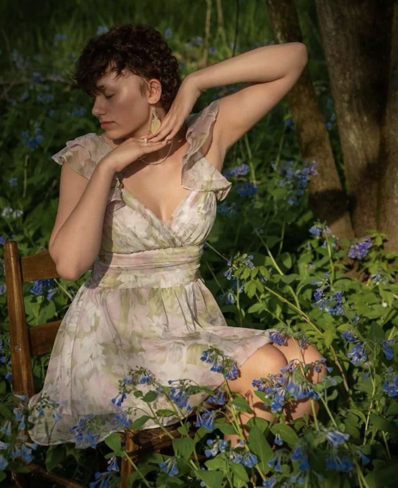 May include: A floral print dress with a V-neck and ruffled sleeves. The dress is light-colored with a pink and green floral pattern. The person is sitting on a wooden chair surrounded by blue flowers and greenery.