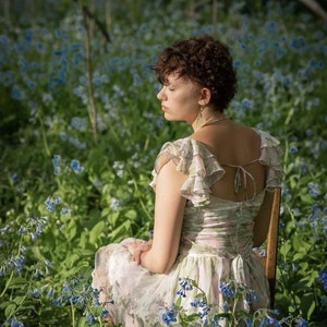 May include: A light-colored floral dress with ruffled sleeves and a tie-back detail. The dress is worn while seated in a field of blue flowers. The dress has a soft, flowing appearance.