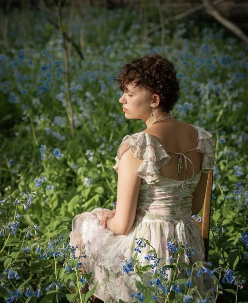 May include: A light-colored floral dress with ruffled sleeves and a tie-back detail. The dress is worn while seated in a field of blue flowers. The dress has a soft, flowing appearance.