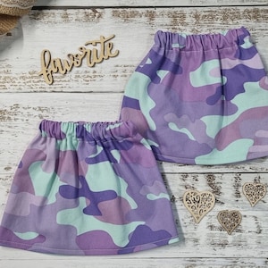 Peut inclure: Deux jupes à motif camouflage violet, lavande et vert menthe avec des tailles élastiquées. Elles sont présentées sur une surface en bois blanc avec des découpes en bois et un ruban de jute.