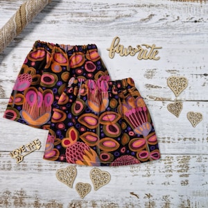 Puede incluir: Dos faldas con cintura elástica, con un colorido estampado floral en tonos rosa, naranja y morado sobre un fondo negro. La palabra "favorite" está escrita en cursiva. También hay recortes de corazón de madera y una flecha decorativa.