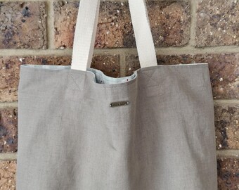 May include: A large, taupe tote bag with cream-colored canvas handles. The bag is made of a sturdy fabric and features a small metal tag near the top. The bag is hanging against a brick wall.
