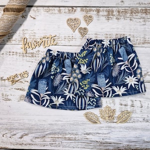 Puede incluir: Dos faldas azul marino con cintura elástica, con un estampado botánico con flores blancas y amarillas y hojas verdes. También se ven recortes de madera con forma de corazón y hoja y la palabra "favorite".