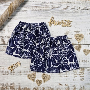 Peut inclure: Deux jupes à imprimé floral bleu marine et blanc avec des tailles élastiques. Les jupes sont décorées de découpes de cœurs en bois et du mot "favorite". Une flèche en bois avec le texte "love this" est également présente.