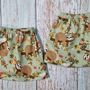 Puede incluir: Dos faldas verde claro con cintura elástica. Las faldas presentan un patrón repetido de perezosos marrones colgados de ramas marrones con flores rosas y amarillas, mariposas y pájaros blancos.