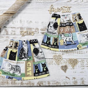 Puede incluir: Dos faldas con temática de gatos con cinturillas elásticas, que presentan un collage de ilustraciones de gatos, texto y patrones en negro, blanco, amarillo y azul. También hay recortes de madera y una cuerda.