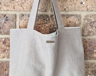 May include: A light beige tote bag with long straps, hanging against a brick wall. The bag has a simple design with a small metal tag that reads "Hand Threads". The interior lining is white with a subtle pattern.