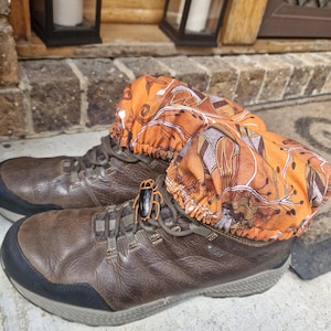 Op de afbeelding: Een paar bruine leren wandelschoenen met oranje stoffen laarsoverschoenen. Een zwart en oranje kevervormige schoenbedel is aan de veters bevestigd. De laarzen staan op een betonnen ondergrond.