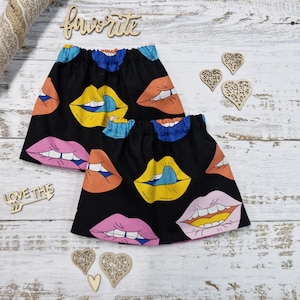 Puede incluir: Dos faldas negras con un estampado de labios coloridos en amarillo, naranja, rosa y azul. Se exhiben sobre una superficie de madera clara, junto con corazones decorativos de madera y las palabras "favorite" y "love this".