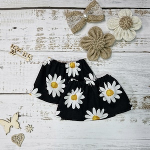 Puede incluir: Faldas negras con estampado de margaritas, con pétalos blancos y centros amarillos. La imagen también presenta lazos y flores de arpillera, además de elementos decorativos de madera. Las faldas son probablemente para niños.