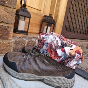 Puede incluir: Una bota de senderismo de cuero marrón con suela negra y una polaina con estampado de gallinas de colores. La polaina presenta un patrón de gallinas en varios colores. La bota está sobre un escalón de hormigón, con faroles y una puerta de madera al fondo.