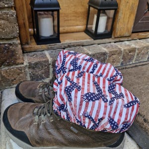 以下が含まれることがあります： 赤、白、青のアメリカ国旗のデザインのブーツカバーが付いた茶色のハイキングブーツ。カバーには、縞模様、星、ワシのシルエットのパターンがあります。背景には、ろうそくが入った2つの黒いランタンがあります。