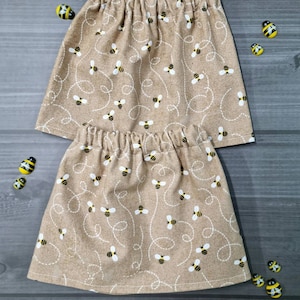 Puede incluir: Dos faldas color beige con cintura elástica, estampadas con líneas punteadas blancas y abejas amarillas y negras. Las faldas se muestran sobre una superficie gris, con adornos en forma de abeja esparcidos.