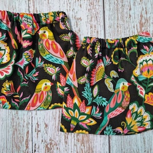Puede incluir: Dos faldas de tela negra con cintura elástica, adornadas con un vibrante estampado floral y de pájaros en amarillo, rosa, turquesa y naranja. Las faldas son probablemente para niños o bebés.
