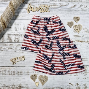 Puede incluir: Dos faldas a juego con un diseño de bandera estadounidense. Las faldas presentan rayas rojas y blancas con siluetas de águilas azules. Las faldas están sobre una superficie de madera blanca con recortes de madera decorativos y la palabra "favorite".