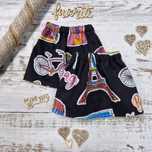 Puede incluir: Dos faldas negras con cinturillas elásticas, con ilustraciones coloridas de una bicicleta, la Torre Eiffel y la palabra "bag". También hay corazones de madera decorativos y la palabra "favorite".