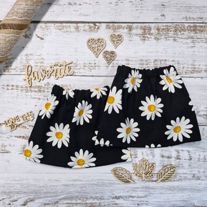 Puede incluir: Dos calentadores de piernas de tela negra con un estampado floral de margaritas blancas y centros amarillos. Los calentadores de piernas se exhiben sobre una superficie de madera blanca con recortes de madera decorativos.