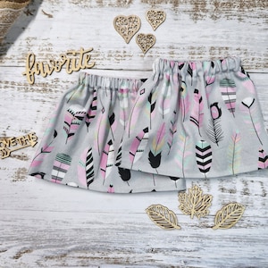 Puede incluir: Dos faldas grises con cintura elástica, adornadas con un estampado de plumas en rosa, negro y turquesa. Recortes de madera de corazones y hojas están dispersos alrededor de las faldas. También está presente la palabra "favorite".