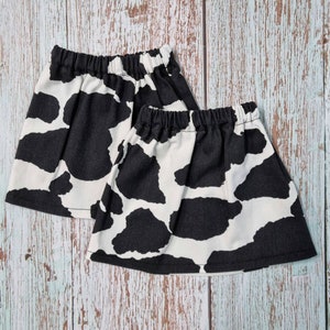Puede incluir: Dos faldas con estampado de vaca en blanco y negro con cinturillas elásticas. Las faldas tienen un diseño acampanado y están hechas de una tela suave. El estampado de vaca es un diseño clásico y divertido.