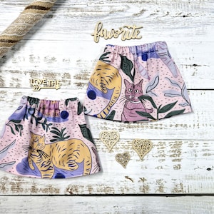 Puede incluir: Dos faldas rosas con temática de gatos con cintura elástica, con un patrón repetido de gatos, plantas y formas abstractas. Las faldas se exhiben sobre una superficie de madera blanca, con recortes de madera que dicen "favorite" y "love this".