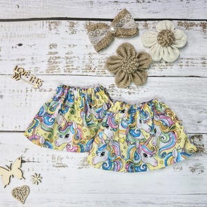 Peut inclure: Deux jupes jaunes avec un motif de licorne, ainsi que des nœuds en toile de jute et des accessoires pour cheveux en forme de fleurs. Les jupes présentent un motif répété de licornes avec des crinières et des queues arc-en-ciel.