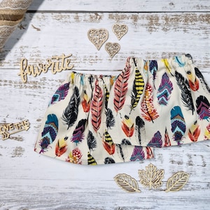 Puede incluir: Dos faldas color crema con cintura elástica, adornadas con un vibrante estampado de plumas en tonos rojos, amarillos, azules y negros. Recortes de madera con forma de corazón y hojas están dispersos.