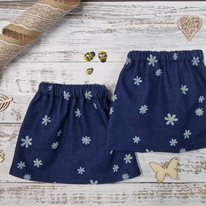 Puede incluir: Dos faldas vaqueras con cinturillas elásticas, adornadas con un estampado de margaritas blancas. Las faldas son de color azul oscuro y se exhiben sobre una superficie de madera clara. También hay recortes decorativos de madera de abejas, una hoja y una mariposa.