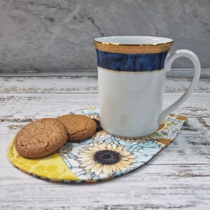Pode incluir: Uma caneca de cerâmica branca com borda dourada e faixa azul, repousando sobre um descanso de copo com estampa de girassol. Dois biscoitos redondos estão no descanso de copo.