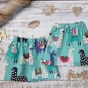 Peut inclure: Deux jupes en tissu turquoise avec une taille élastique, ornées de lamas colorés. Les jupes sont présentées sur une surface en bois blanc, avec des découpes en bois décoratives et un ruban de jute.