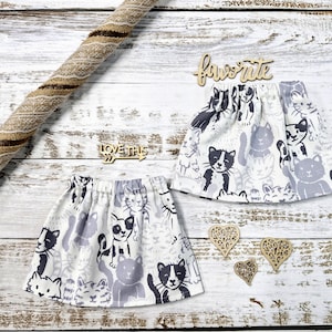Puede incluir: Dos faldas con estampado de gatos con cintura elástica, con un patrón repetido de gatos negros, blancos y grises. Recortes de madera deletrean "favorite" y "love this". Una cuerda decorativa está en el fondo.