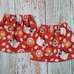 Puede incluir: Dos faldas rojas con cintura elástica, con un estampado repetido de koalas blancos, girasoles y hojas. Las faldas están hechas de una tela ligera, perfectas para el clima cálido.