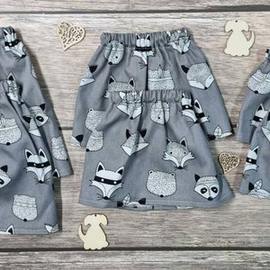 Puede incluir: Tres faldas grises con cintura elástica, cada una adornada con un patrón repetido de zorros y mapaches en blanco y negro. Las faldas se exhiben sobre una superficie de madera, con recortes decorativos de madera de corazones y perros.