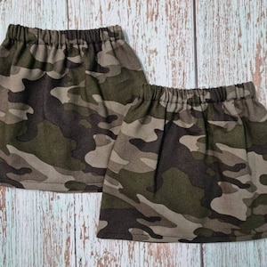 Puede incluir: Dos faldas con estampado de camuflaje. Cuentan con cintura elástica y un diseño corto y acampanado. La tela presenta una mezcla de colores verde, marrón y beige, creando una estética de inspiración militar. Las faldas se exhiben sobre una superficie de madera clara.