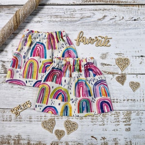 Peut inclure: Deux jupes blanches avec un motif arc-en-ciel à l'aquarelle. Les jupes ont une taille élastique et sont décorées d'arcs-en-ciel colorés. Des découpes en bois de cœurs et le mot "favorite" sont également présentes.