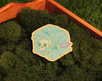 May include: A sticker featuring a light blue cube design with a skull, bones, and a sword. The sticker has a yellow border and is set against a background of green moss inside a wooden tray.