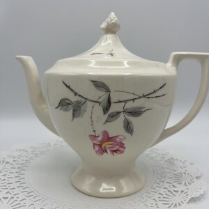 May include: White teapot with a floral design featuring pink roses and gray leaves. The teapot has a lid with a knob on top and a spout on the side.