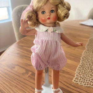 Puede incluir: Una muñeca de porcelana vintage con cabello rubio rizado, vestida con un vestido a rayas rosa y blanco y sandalias blancas. La muñeca está de pie sobre un soporte blanco.
