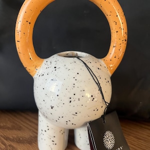 Pode incluir: Um vaso de cerâmica em forma de kettlebell, com um corpo branco salpicado e uma alça laranja. A alça e o corpo são salpicados com pontos pretos. O vaso tem três pernas curtas e uma etiqueta "LIVETRENDS".