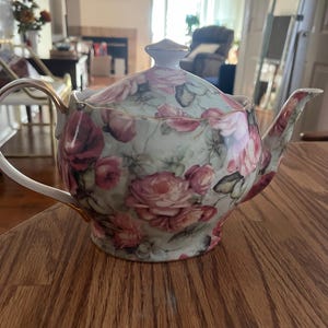 May include: A white porcelain teapot with a floral pattern of pink roses and butterflies. The teapot has a gold trim around the lid and spout.
