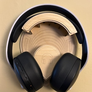 Puede incluir: Un auricular blanco y negro está colgado de un soporte de madera con un icono de auriculares. El soporte está diseñado para mantener los auriculares en su lugar.
