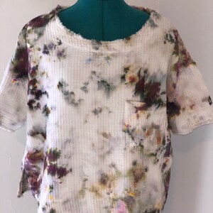 May include: A short-sleeved, off-white shirt with a pocket, featuring a tie-dye pattern in shades of purple, green, and yellow. The shirt has a ribbed texture and a raw, frayed edge around the neckline and sleeves.
