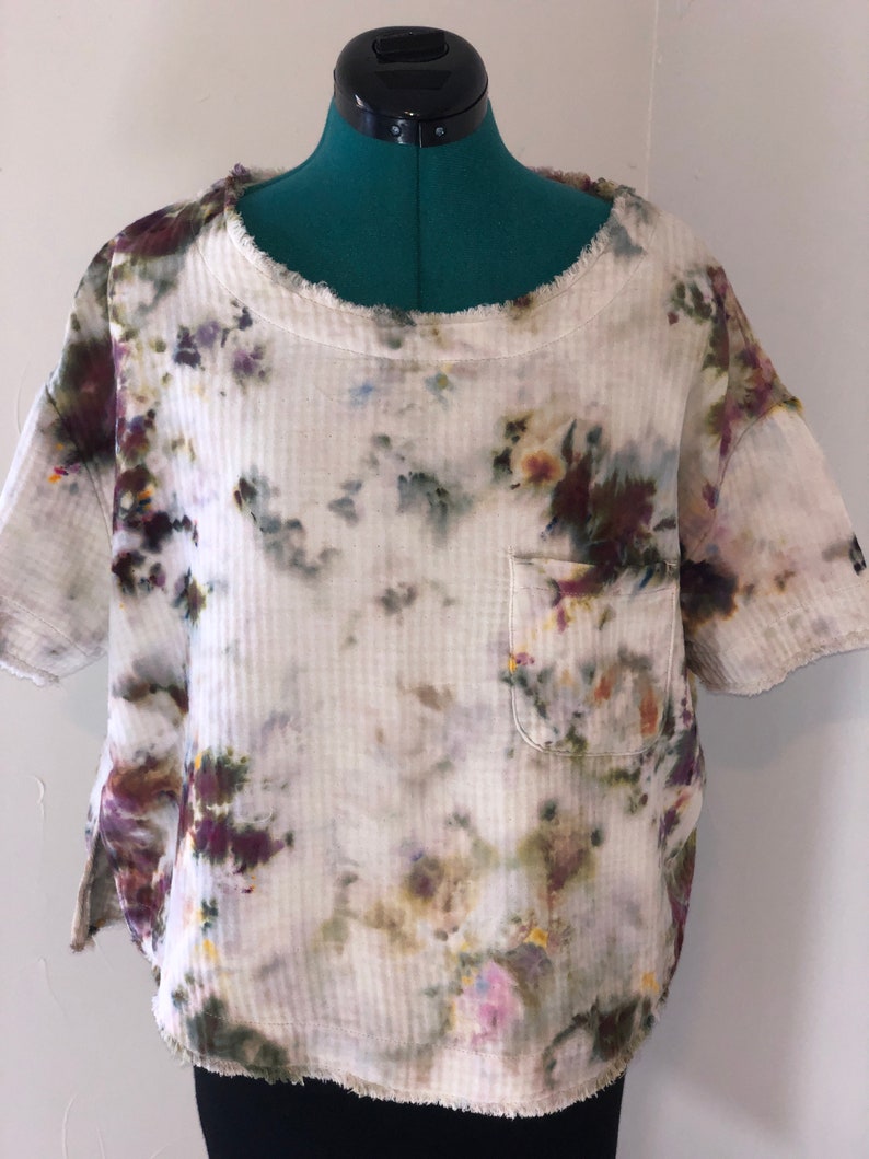 May include: A short-sleeved, off-white shirt with a pocket, featuring a tie-dye pattern in shades of purple, green, and yellow. The shirt has a ribbed texture and a raw, frayed edge around the neckline and sleeves.