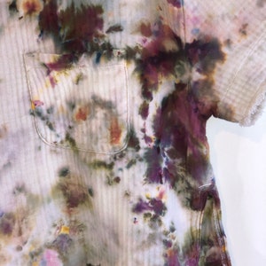 May include: A short-sleeved shirt with a pocket, featuring a unique tie-dye pattern in shades of purple, green, and yellow on a cream-colored fabric. The shirt has a textured, striped design and frayed edges.