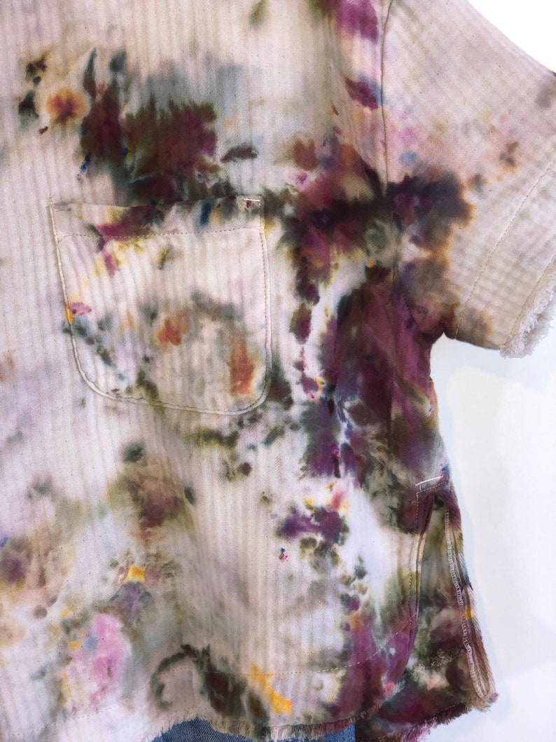 May include: A short-sleeved shirt with a pocket, featuring a unique tie-dye pattern in shades of purple, green, and yellow on a cream-colored fabric. The shirt has a textured, striped design and frayed edges.