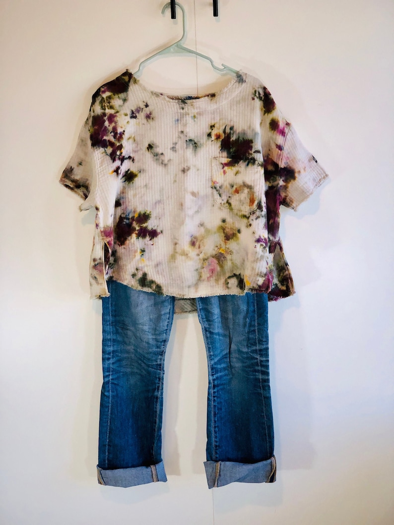 May include: A tie-dye short-sleeve shirt with a pocket, paired with blue jeans. The shirt features a white base with colorful floral-like patterns in shades of purple, green, and yellow. The jeans are cuffed at the ankles.