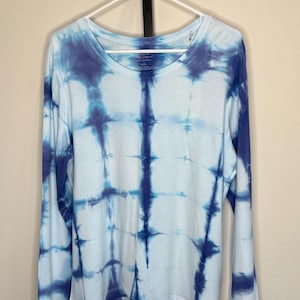 Może przedstawiać: Koszulka z długim rękawem w stylu tie-dye w odcieniach niebieskiego i białego wisi na białym wieszaku. Koszulka ma wzór kratki stworzony techniką tie-dye. Tkanina wydaje się miękka i lekka, odpowiednia do noszenia na co dzień.