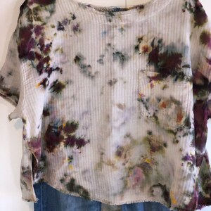 May include: A tie-dye short-sleeve shirt with a pocket, displayed on a hanger. The shirt features a ribbed texture and a mix of colors, including purple, green, and yellow, against a white background. Frayed edges add a casual touch. Paired with blue jeans.